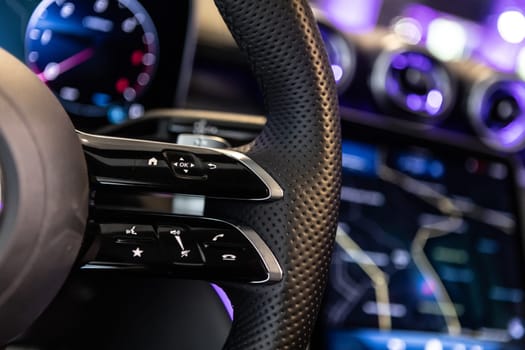 Close-up shot of a luxury car steering wheel featuring control buttons and sleek leather details, with a blurred background displaying a high-tech dashboard and navigation screen.