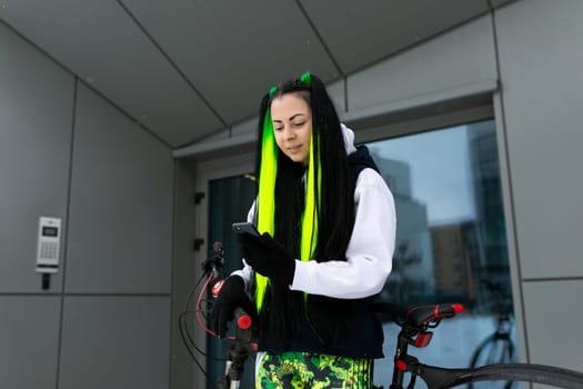 A woman with dyed green hair is standing outdoors, holding a bicycle. She is looking at the camera with a confident expression, showcasing her unique style and transportation choice.