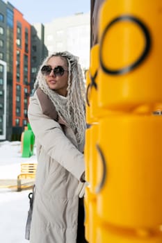 A woman is standing next to a yellow pole on a sidewalk. She appears to be waiting or holding onto the pole. The background shows a cityscape with buildings.
