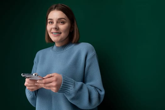 A woman wearing a blue sweater is seen holding a cell phone in her hand. She appears to be using the phone for communication or browsing. The background is simple and non-distracting.
