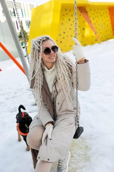 A woman sits on a swing covered in snow, bundled up in warm clothing. The snow-covered ground contrasts with her dark outfit as she looks contemplative.