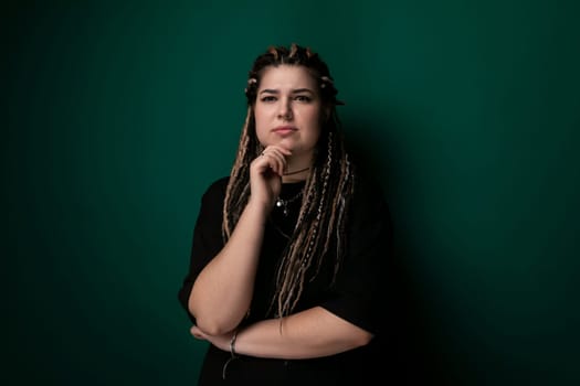A woman with dreadlocks is leaning against a vibrant green wall. She appears relaxed and casual, with a confident posture. The contrast between her hair and the wall adds visual interest to the scene.