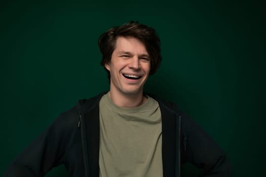 A man is standing in front of a vibrant green wall, smiling brightly. His expression exudes happiness as he poses for the camera in a cheerful manner.