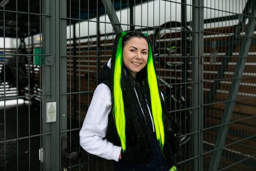 A woman with green hair is standing in front of a wooden fence. She appears to be looking off into the distance. The fence is weathered and has peeling paint. The woman is wearing a casual outfit and seems relaxed.