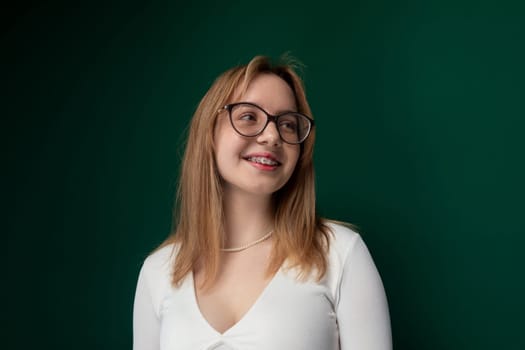 A woman with glasses is standing in front of a vibrant green wall. She appears to be looking straight ahead, with a neutral expression on her face.
