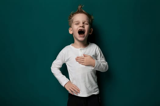A young male child is standing in front of a solid green wall. He appears to be looking straight ahead, with a neutral expression on his face. The wall provides a simple backdrop to the scene.