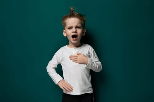 A young boy wearing a blue t-shirt and jeans is standing in front of a bright green wall. He has short brown hair and appears to be looking slightly off to the side.
