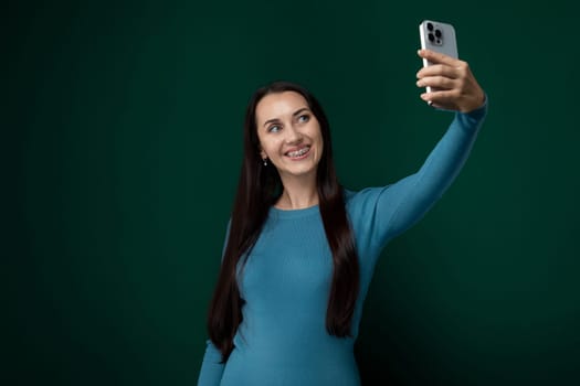 A woman is holding her cell phone in front of her face, smiling as she takes a selfie. She appears to be standing outdoors.
