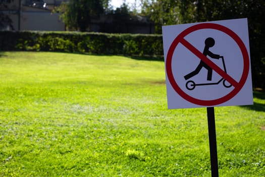 Sign in grassy field says no scooter in park. Sign is red and black. Symbol prohibiting the use of scooter near field green in background