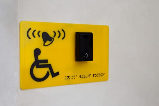 Yellow sign on wall of building with sign of person in wheelchair next to button to call help and braille text for blind people. Helping people with disabilities on the street in city, accessible environment