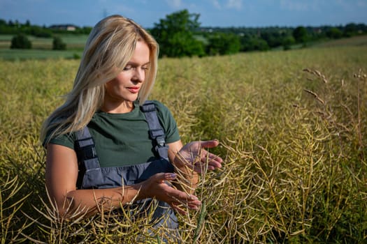 A female farmer in work overalls gently holds a rapeseed plant, checking the maturity of its green and yellowing pods. A rural village and partly cloudy blue sky provide a serene backdrop to the scene.