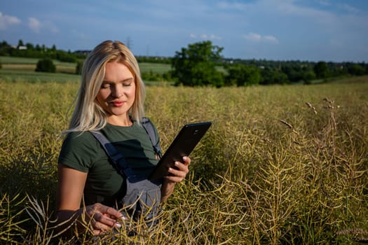 A woman farmer is checking the quality of rapeseed on the middle of the field and holding a tablet. She is checking the information about her harvest. She owns a rapeseed field and is checking this year's harvest. She is an experienced farmer, caring for her crops in a vast field. She has blonde hair and smiles. She is pleased and satisfied with her yields. The soil is fertile and productive. The rapeseed is yellow and ready for harvest. Satisfied with the bountiful and lavish harvest, the woman farmer, adorned in working clothes and with sun-kissed skin, gazes upon the expansive yellow landscape with a sense of fulfillment. With the blue sky adorned with fluffy white clouds overhead, she stands in profile, her spirit merging with the warmth of the sun, celebrating the culmination of her dedication and the richness of the land's yield.