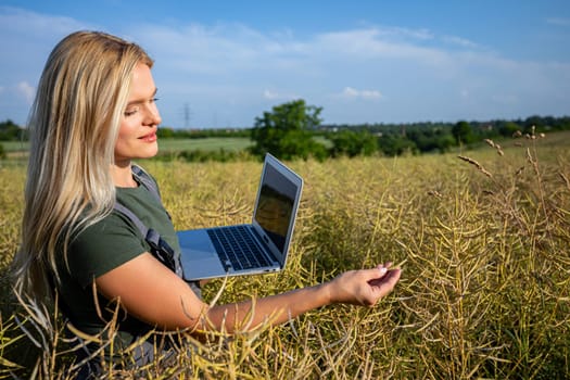 A woman farmer is checking the information about rapeseed using a laptop. She is gently touching the rapeseed and checking if everything is ok with her harvest. She is checking the information about her harvest. Farmer owns a rapeseed field and is checking this year's crop. She has blonde hair and she stands in profile. The rapeseed is yellow and ready for harvest. Satisfied with the bountiful and lavish harvest, the woman farmer, adorned in working clothes and with tanned skin, gazes upon the expansive yellow landscape with a sense of fulfillment. The sky is blue adorned with fluffy white clouds overhead. The day is warm and sunny.