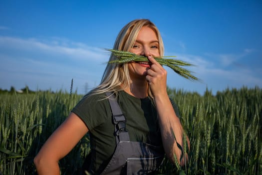 In a picturesque wheat field, a female farmer stands, her one hand cradling wheat stalks. She playfully arranges them near her face, creating a whimsical mustache-like effect or whiskers, while making a silly face. Her expression reflects her delight in the bountiful harvest. With sun-kissed skin and blonde hair, clad in work attire, she embodies the essence of rural life. The vibrant green color of the wheat stalks contrasts beautifully against the golden backdrop of the field. Amidst the rolling waves of grain, she stands, a symbol of agricultural pride and joy. This image captures the lighthearted spirit of farming, where hard work meets moments of playful enjoyment. Against the backdrop of the expansive wheat field, her contentment shines through, a testament to the rewards of agricultural labor. With each stalk held tenderly, she celebrates the abundance of nature's bounty and the timeless connection between farmer and land
