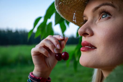 The close-up image captures the essence of a woman's face and hands, revealing her painted, full lips. She is holding red cherries in her hand. Her gaze is directed towards the sky, framed by a backdrop of a cherry tree branch and a blurred forest in the distance. Imperfections adorn her skin, adding a raw authenticity to her presence, while her piercing blue eyes reflect a sense of introspection. Against her wrist, a colorful bracelet crafted from beads adds a playful touch to her demeanor. She has straw hat on her head. The cherries in her grasp exude ripeness, promising a burst of sweetness with every bite. Amidst the tranquil surroundings of the orchard, she stands as a symbol of connection to nature, her contemplative gaze hinting at a moment of quiet reflection amidst the beauty of the natural world