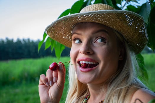 In the close-up shot, the woman's face and hands take center stage, her lips painted and full, complementing her vibrant blue eyes and playful smile. With an air of contentment, she eagerly anticipates indulging in the ripe cherries. In the background, a cherry tree branch and a blurred forest add to the serene atmosphere. Holding a single red cherry between her teeth, the stem protruding, she reveals her white teeth and tongue. The cherries, vibrant red and fully ripe, promise a burst of flavor. Completing her ensemble is a straw hat atop her head, perfect for the pleasant summer day. Her carefree smile and playful gaze add a sense of whimsy to the scene, as she looks off to the side with amusement. In this moment of simple joy, she embodies the essence of summertime bliss, basking in the warmth of the sun and the delight of nature's bounty.