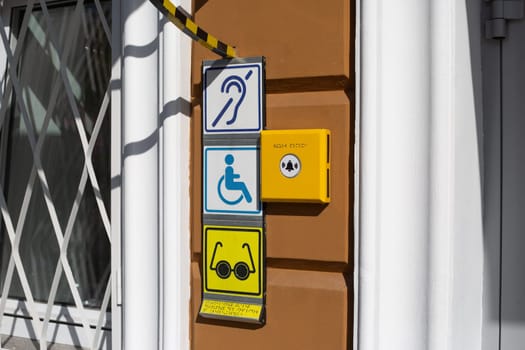 Sign with international sign of person with disabilities in wheelchair on wall near entrance to building. Deaf and blind person symbol with braille text