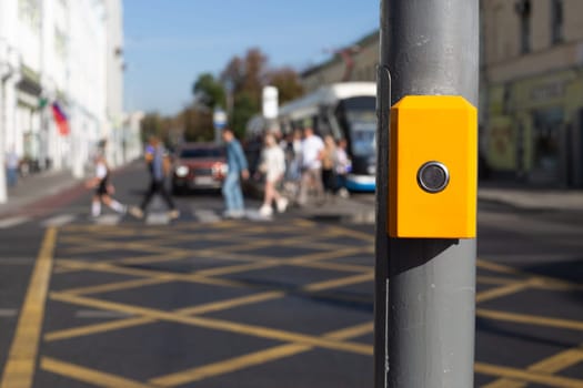 Button for changing traffic light signal for pedestrians to cross an intersection. Control panel for waiting to cross the road hangs on pole on the background of people cross road