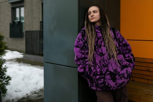 Stylish informal woman with dreadlocks dressed in a purple jacket walks through the city in winter.