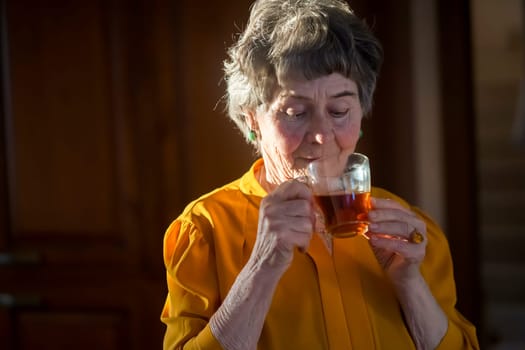 An elderly well-groomed gray-haired woman holds a glass cup of tea in her hands and enjoys its taste, the grandmother has a good time in her cozy house, relaxing in the kitchen in the rays of the sun.