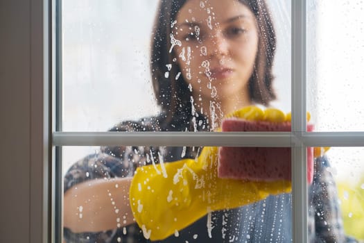 A young girl in yellow rubber gloves and a uniform washes the windows thoroughly with a detergent, wiping them dry with a rag, the woman is cleaning the house.