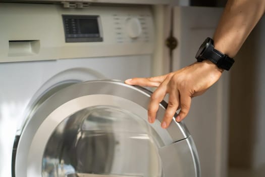 A male hand opens the door of a new washing machine in his modern, bright bathroom, close-up. The man washes clothes in the laundry.