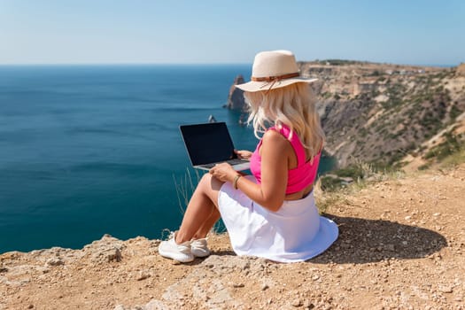 A woman is sitting on a rocky beach with a laptop open in front of her. She is wearing a pink top and a straw hat. Concept of relaxation and leisure