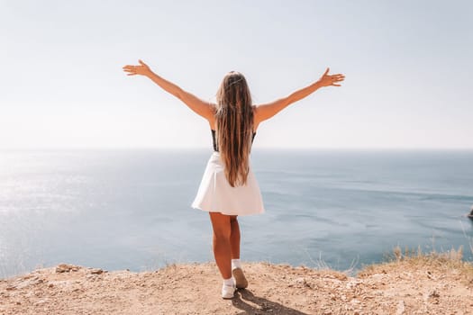 A woman stands on a rocky beach with her arms raised in the air. The sky is clear and the ocean is calm. The woman is happy and enjoying the beautiful scenery