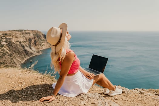 A woman is sitting on the beach with a laptop open in front of her. She is wearing a pink top and a straw hat. Concept of relaxation and leisure