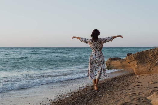 A woman in a floral dress stands on a sandy beach, arms outstretched, facing the ocean. The waves crash against the shore.