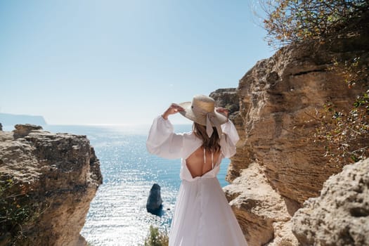 woman white dress is standing on a cliff overlooking the ocean. She is wearing a straw hat and she is enjoying the view