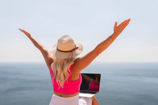 A woman wearing a pink top and hat is holding a laptop and looking out at the ocean. She is enjoying the view and the technology she is using