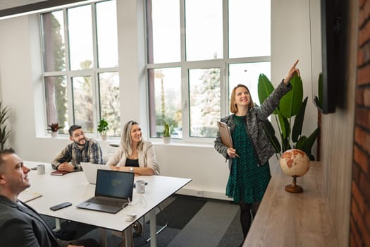 In the picture, a woman is confidently giving a presentation to a diverse group of people. She gestures towards a screen displaying charts and graphs, engaging her audience with her speech.