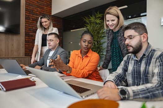 A diverse group of individuals is gathered around a large table, each focused on their laptop screens. The atmosphere is productive and collaborative as they engage in discussions and work together on various projects.