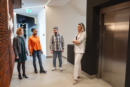A diverse group of individuals of varying ages and genders are standing together in a hallway, appearing to engage in conversation or waiting for something. The hallway is well-lit and lined with doors on either side.