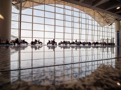 Passengers sitting in chairs in a line are waiting to board their flights in a modern airport terminal