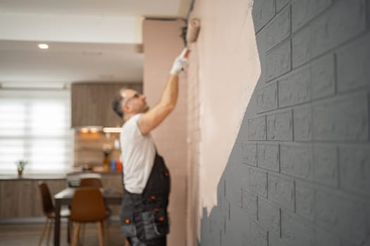 A man is using a paint roller to coat a brick wall with fresh paint. He is focused on his task, ensuring even coverage on the surface.