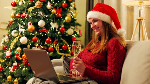 A woman in a red sweater and Santa hat holding a glass of wine while using a laptop next to a decorated Christmas tree. Indoor setting with festive holiday decor