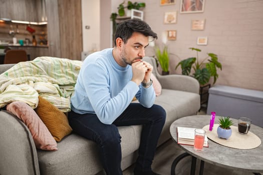 Brown-haired depressed man is sitting on the couch in the living room. He is pensive and worried.
