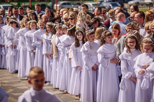 Group of children wearing white clothes standing in line during their first holy communion ceremony with their families in the background