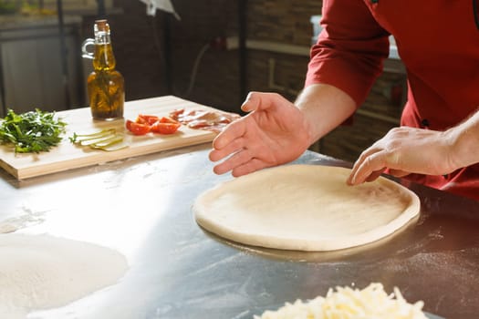 A person shaping pizza dough on a floured surface in a kitchen, with ingredients like tomatoes and pickles in the background.