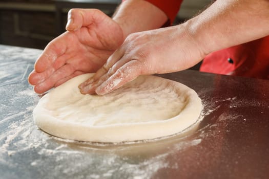 A person shaping pizza dough on a floured surface in a kitchen, with ingredients like tomatoes and pickles in the background.