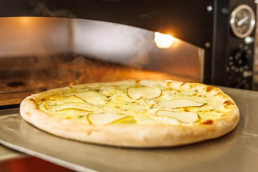 A freshly baked pizza with thin slices of pear on top, placed on a metal tray. The pizza is in a wood-fired oven, with steam rising in the background, creating a warm and inviting atmosphere.