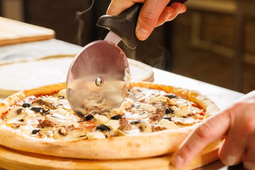 Close-up of a person cutting a freshly baked pizza with a pizza cutter. The pizza has toppings including mushrooms, olives, and cheese.