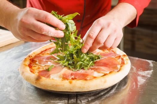 A chef adding fresh arugula to a pizza with prosciutto on a floured surface.