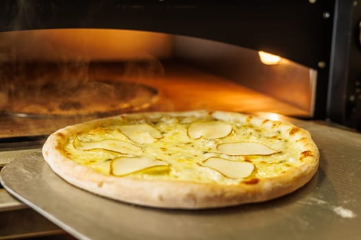 A freshly baked pizza with thin slices of pear on top, placed on a metal tray. The pizza is in a wood-fired oven, with steam rising in the background, creating a warm and inviting atmosphere.