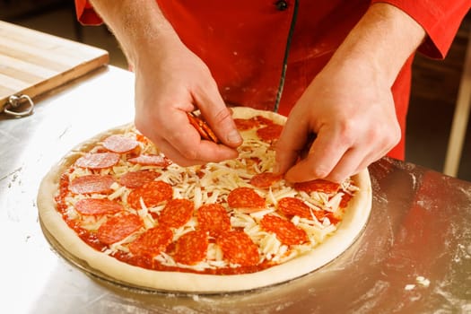 A person in a red shirt preparing a pepperoni pizza by placing pepperoni slices on top of cheese and tomato sauce.