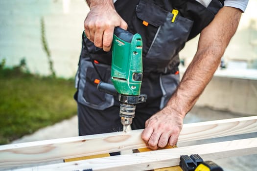 Unrecognizable male carpenter using electric drill on a wooden plank outdoors.