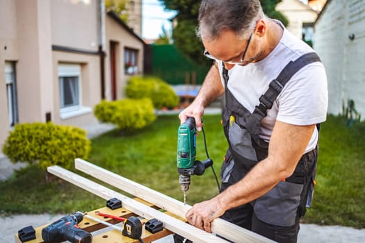 Mature male carpenter using electric drill on a wooden plank outdoors.