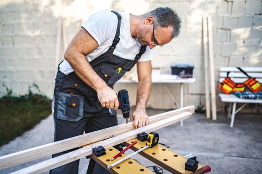 Mature male carpenter using electric drill on a wooden plank outdoors.